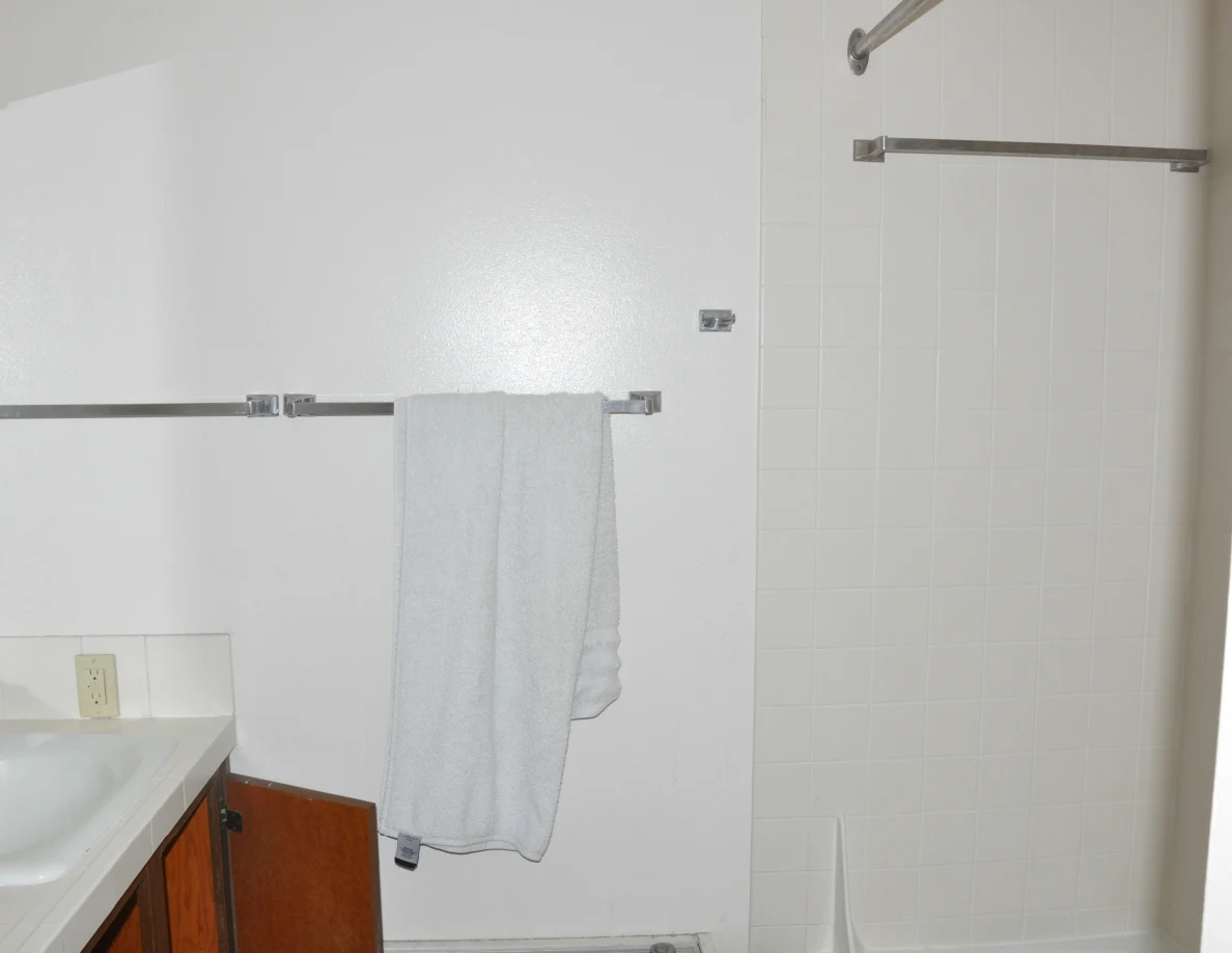 Bathroom wall with a hanging gray towel beside tiled bathtub walls and an open vanity cupboard.