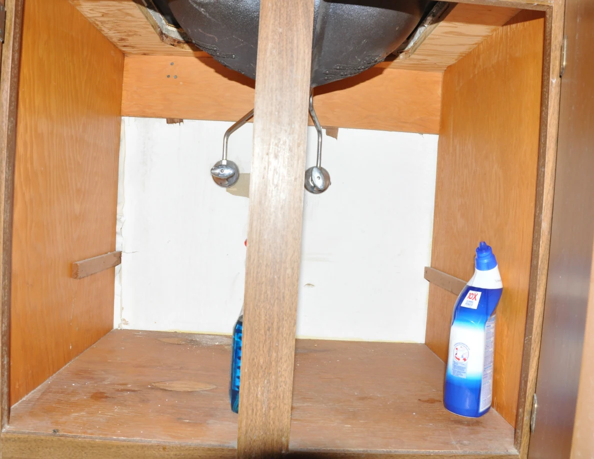 Under-sink cupboard with exposed valves and cleaning bottles on its wooden base.