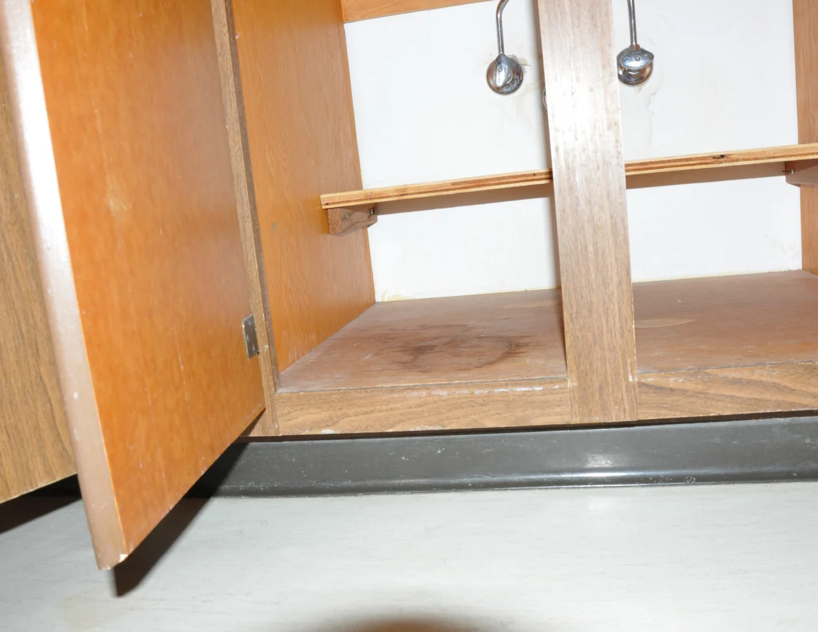 Empty open under-sink cupboard with valves, a narrow shelf and a circular stain on the base.