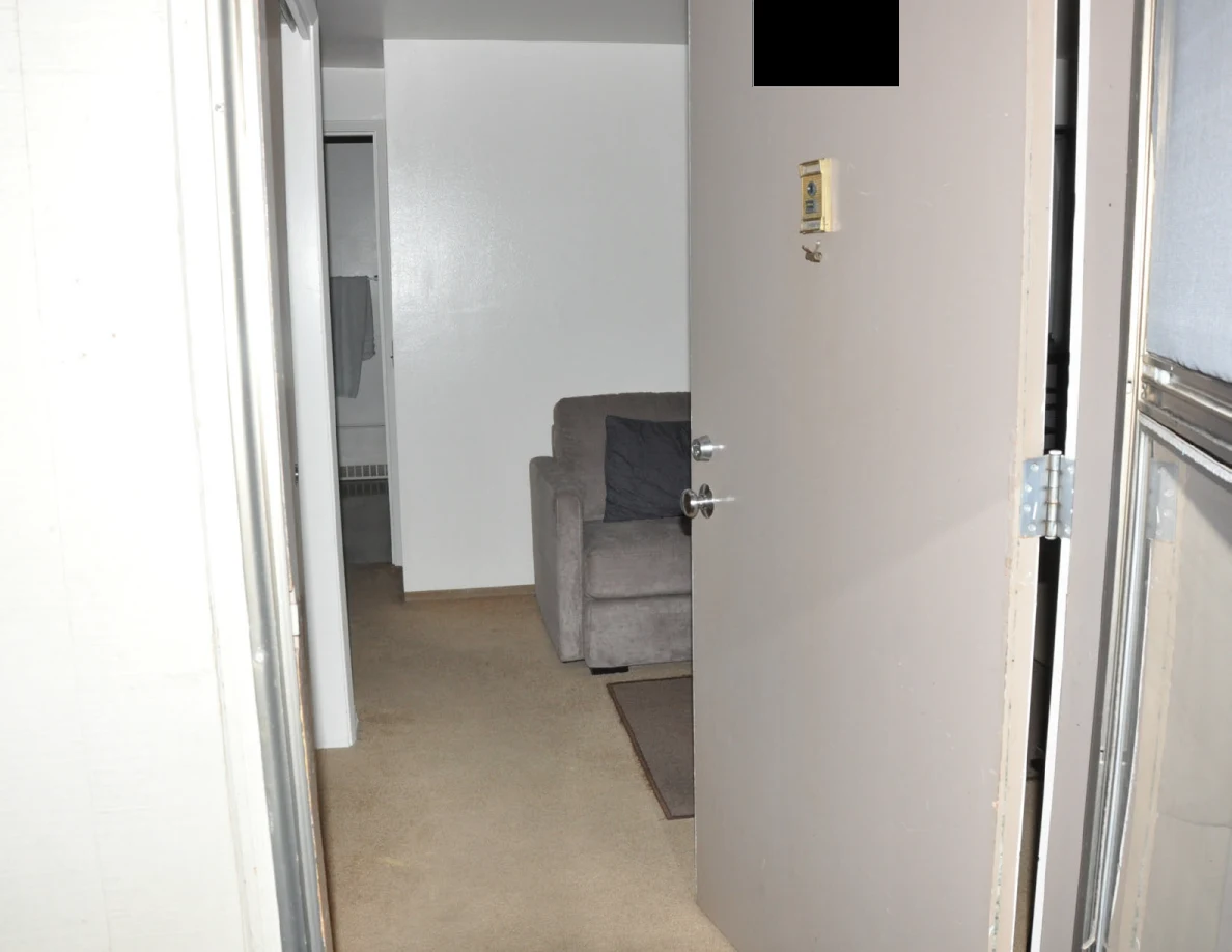 Open entrance door with a black redaction, showing a gray armchair and hallway beyond.