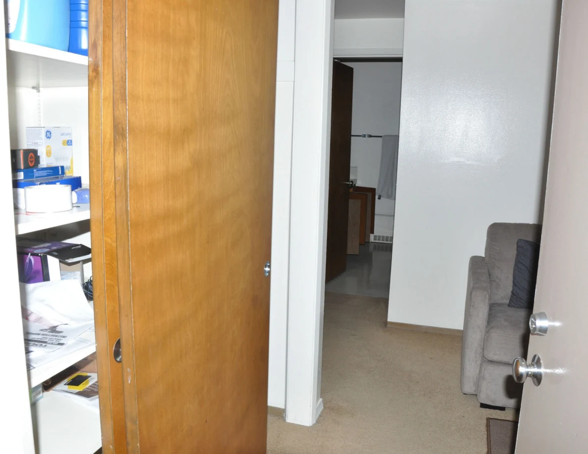 Open storage cupboard with bottles, boxes and paper stacks beside a carpeted hallway.