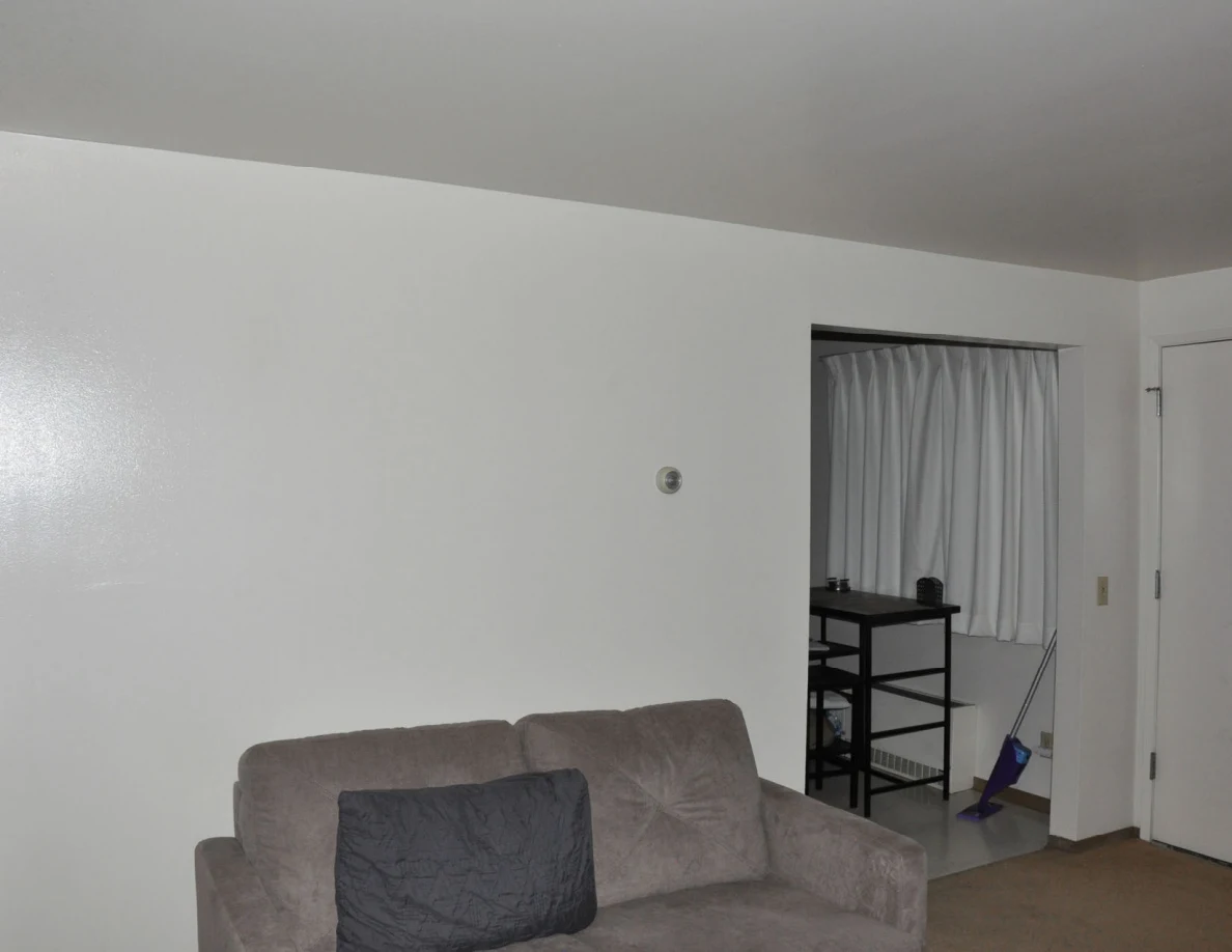 Gray loveseat below a plain wall, with a small dining area and broom visible beyond.