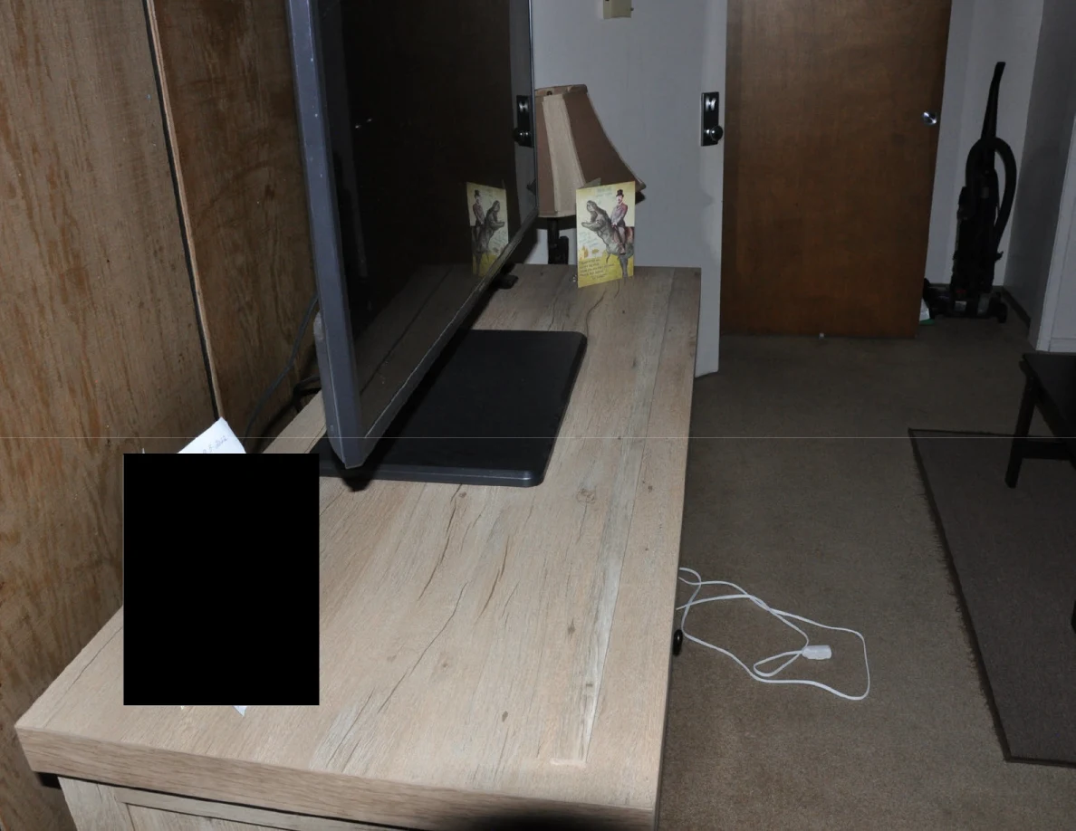 Side view of a television cabinet with a black redaction, and white cords on the carpet.
