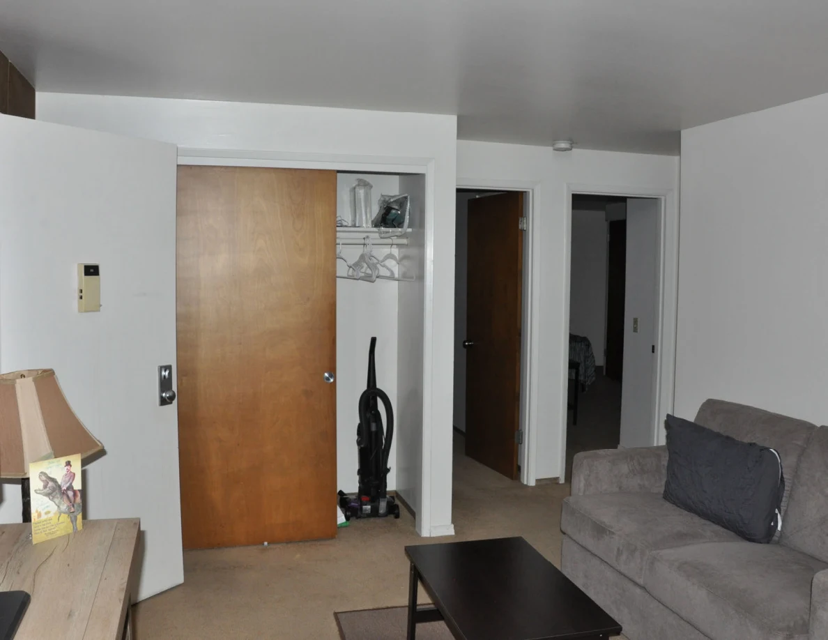 Living room with gray loveseat, coffee table, lamp and open closet holding a vacuum.