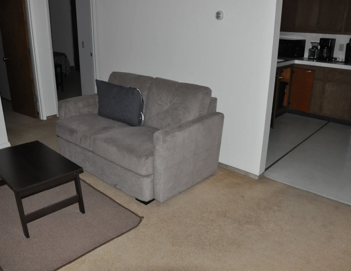 Gray loveseat beside an open kitchen entrance with wooden cupboards.