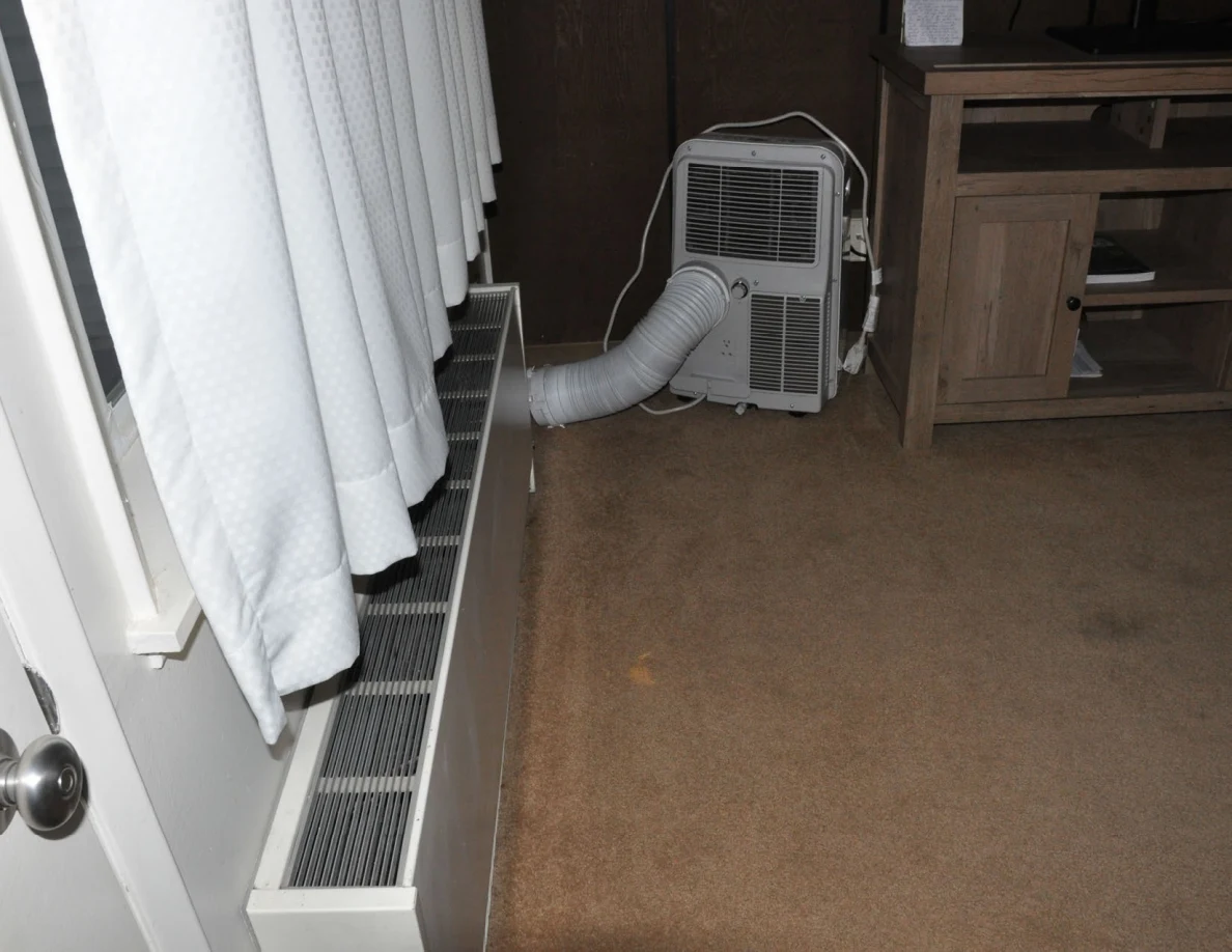 Portable air conditioner with exhaust hose beside curtains, wall heater and television cabinet.