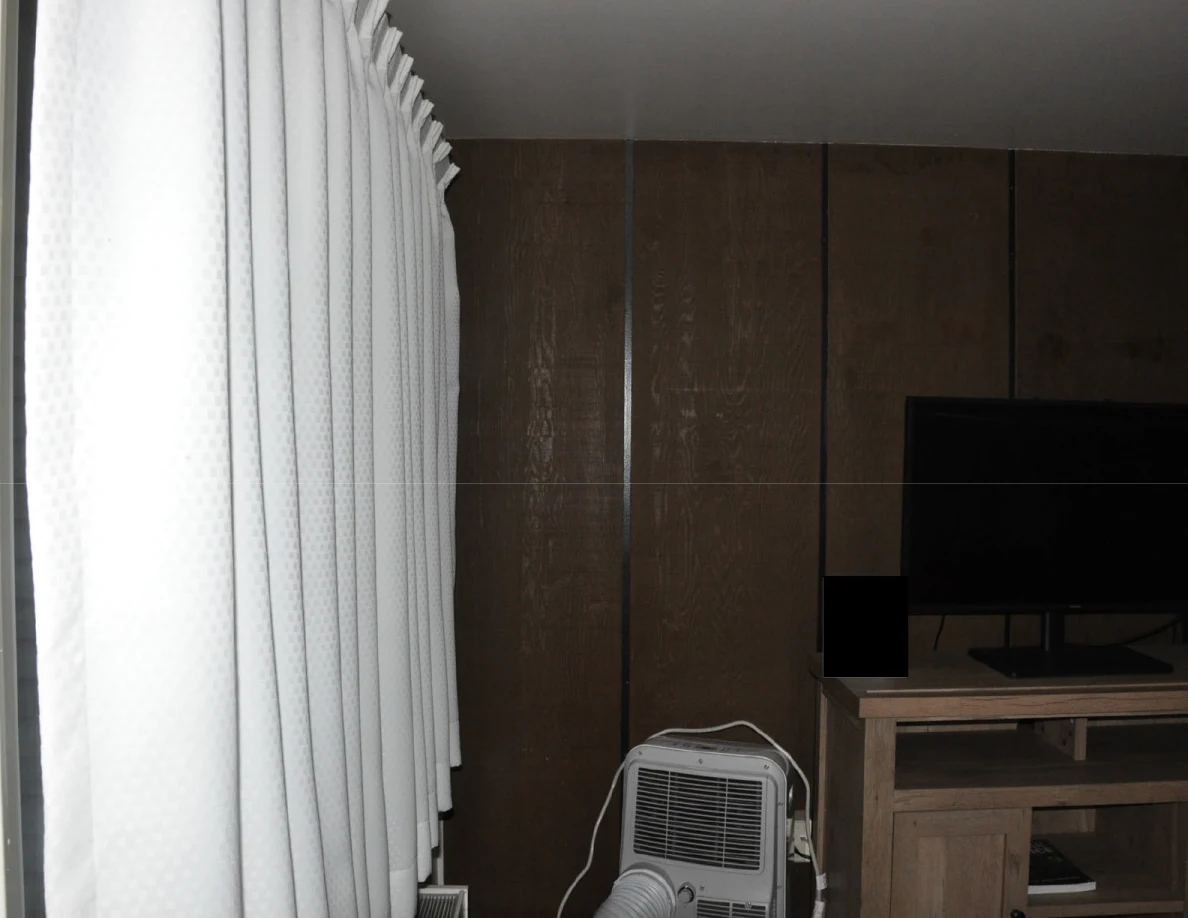 White curtains beside a portable air conditioner and television against wood paneling.