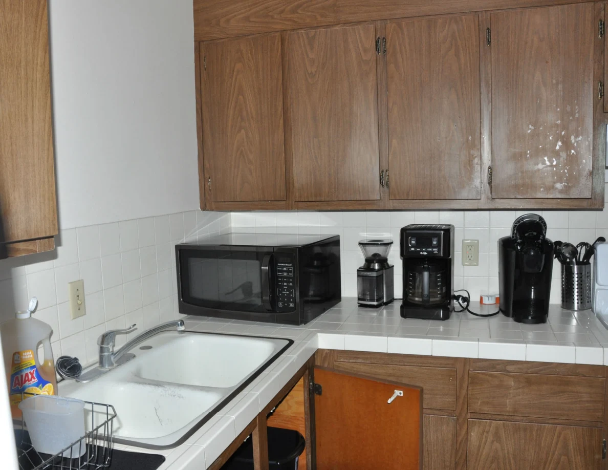 Kitchen double sink and counter with microwave, coffee makers and utensil holder beneath wooden cupboards.