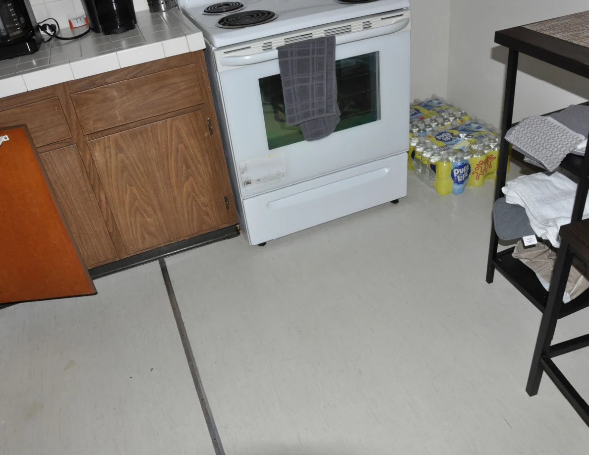 Lower kitchen view with white oven, wooden cupboard and packs of canned drinks beside a table.