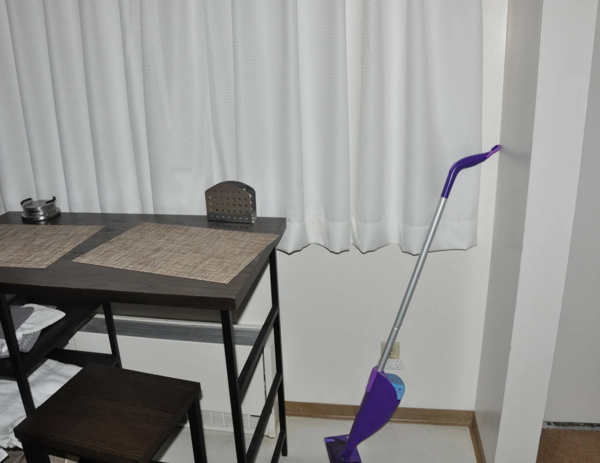 Small dining table with placemats and a purple mop beside closed white curtains.