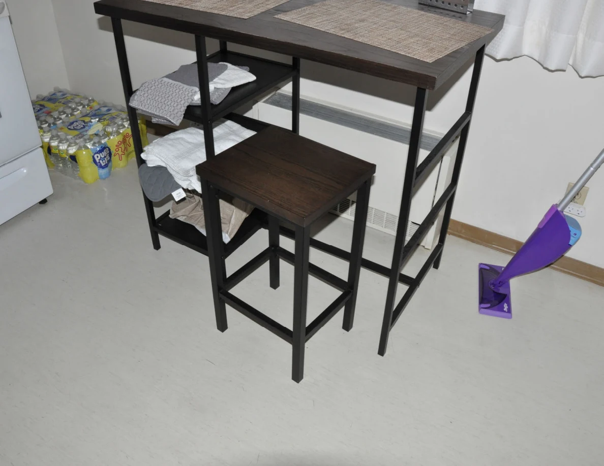 Stools under a tall table with fabric on shelves, beside packs of canned drinks and a mop.