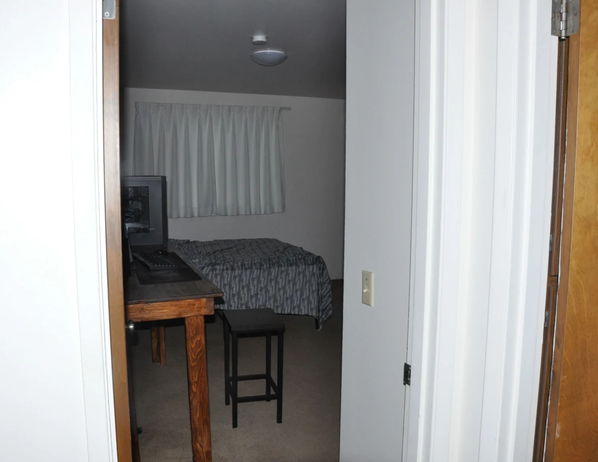 Bedroom entrance showing a desk, computer equipment, black stool and bed beneath curtains.