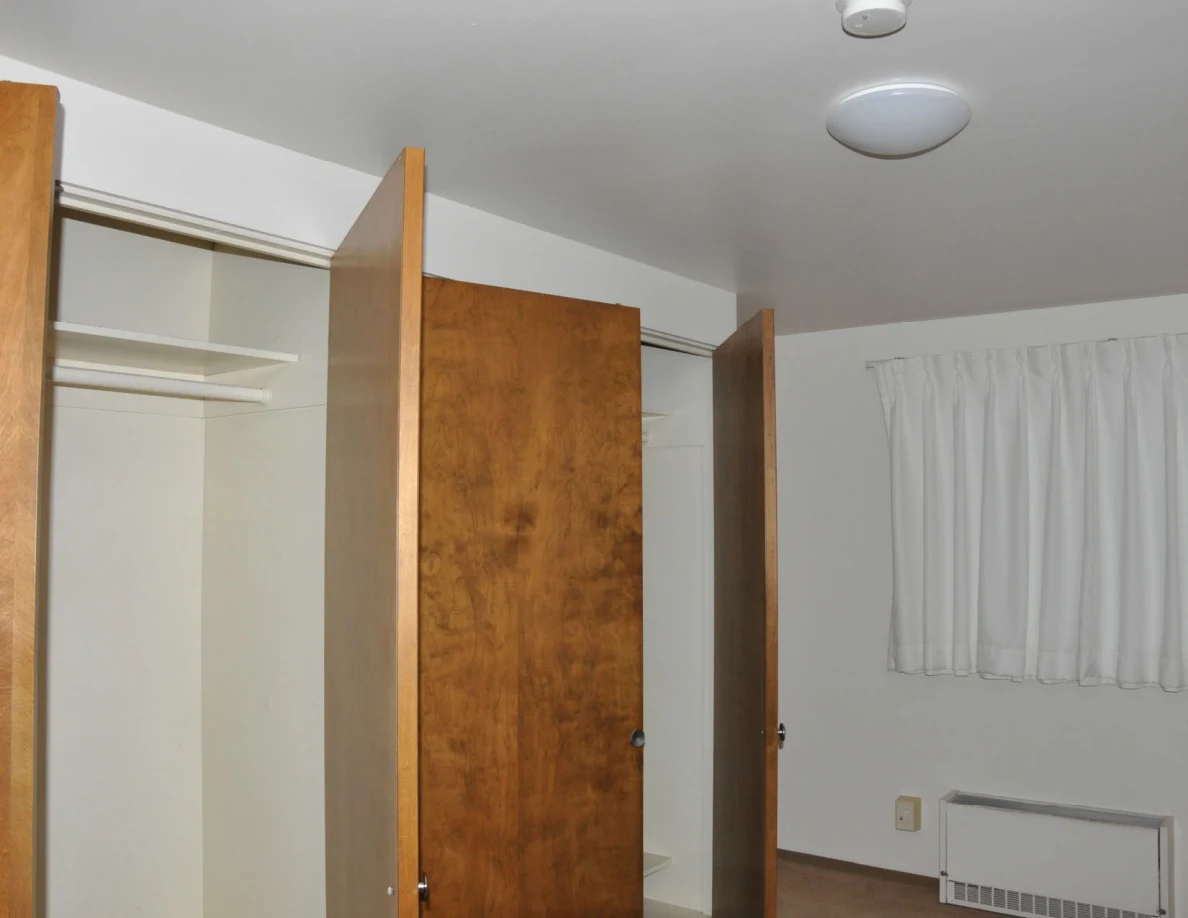 Open empty closets beside white curtains, with a ceiling light and smoke alarm overhead.