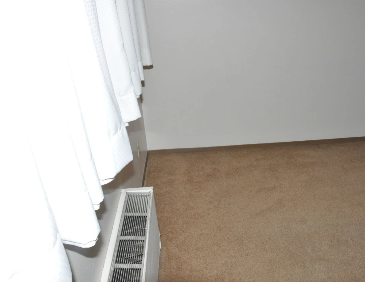 Beige carpet beside a wall heater and closed white curtains.