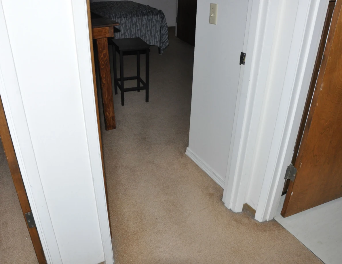 Bedroom doorway with a desk, tall stool and bed visible beyond.