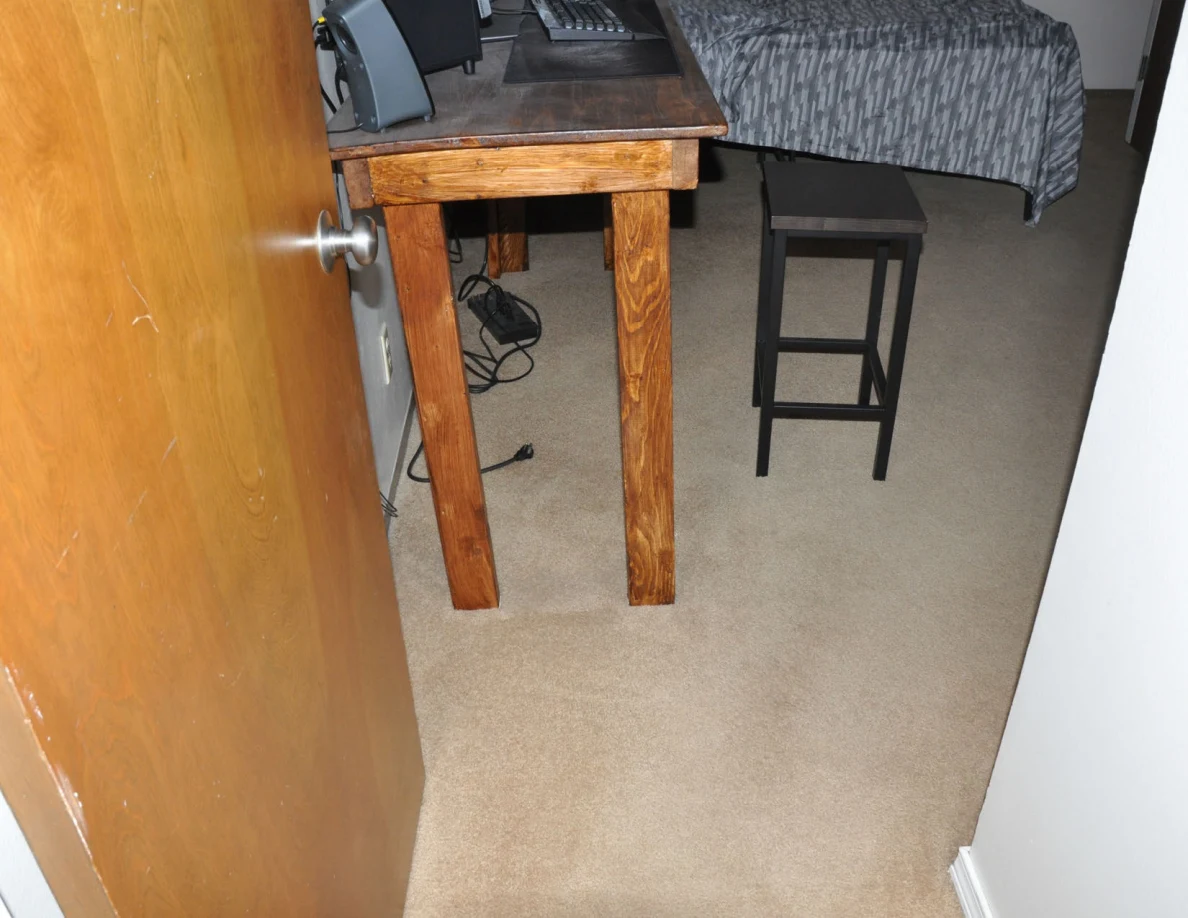 Wooden computer desk and black stool on carpet, with a bed behind.