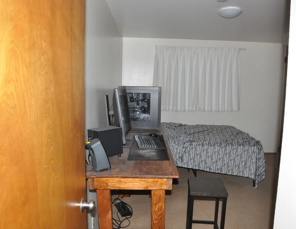 Bedroom with a desktop computer, wooden desk, stool and gray-patterned bedspread.