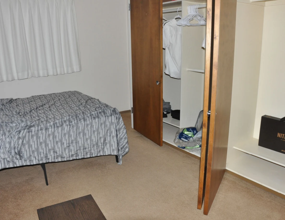 Gray-patterned bedspread beside open closets with shirts and packaged bedding.