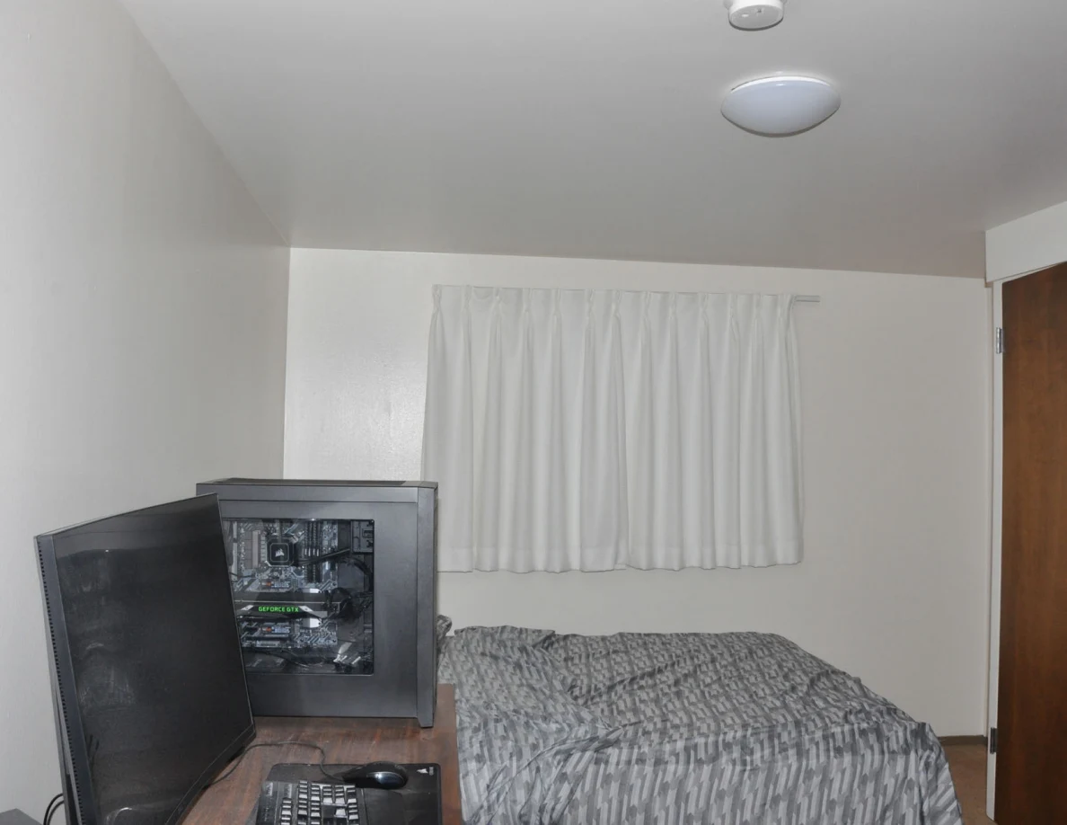 Computer tower and monitor on a desk beside a bed beneath white curtains.
