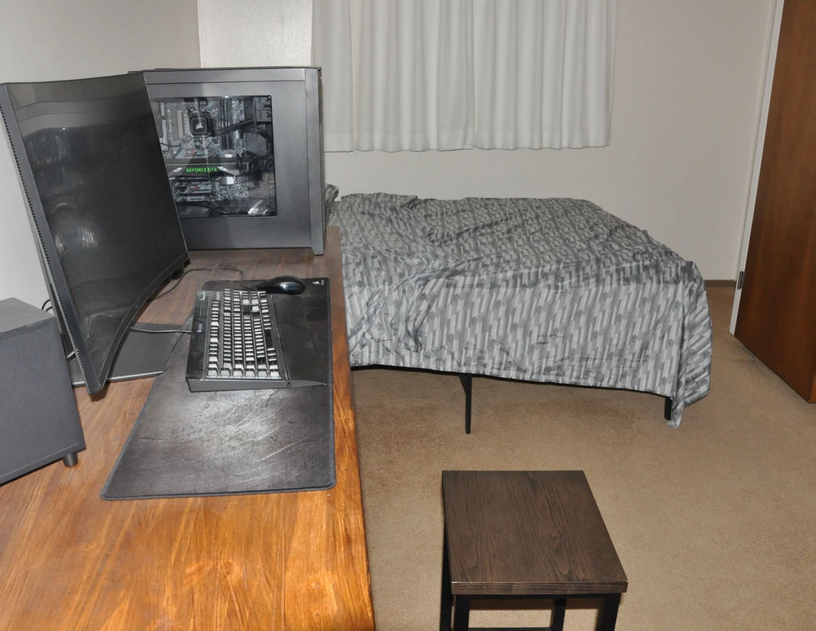 Wooden computer desk with monitor, keyboard and tower beside a bed and black stool.