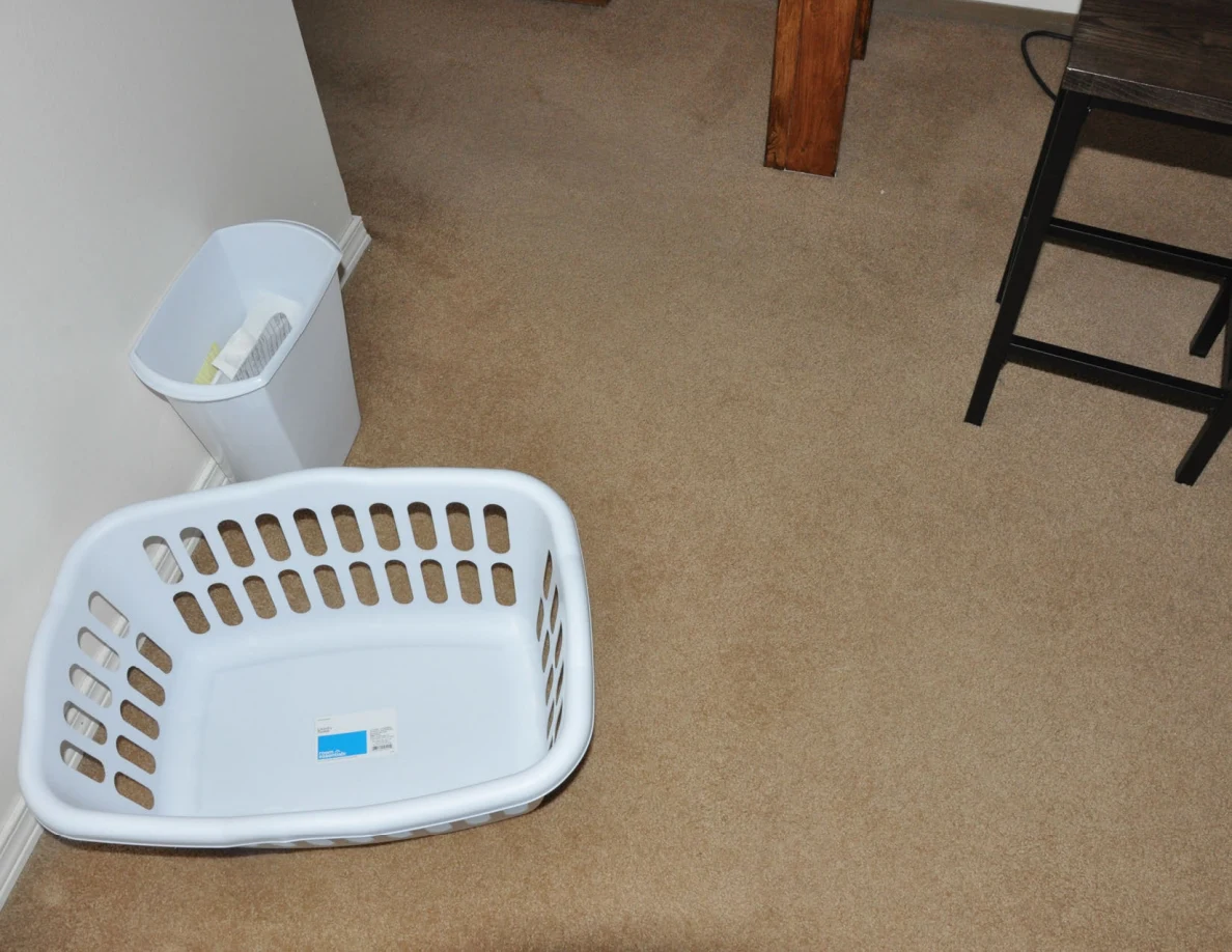White laundry basket and wastebasket beside a desk leg and black stool.