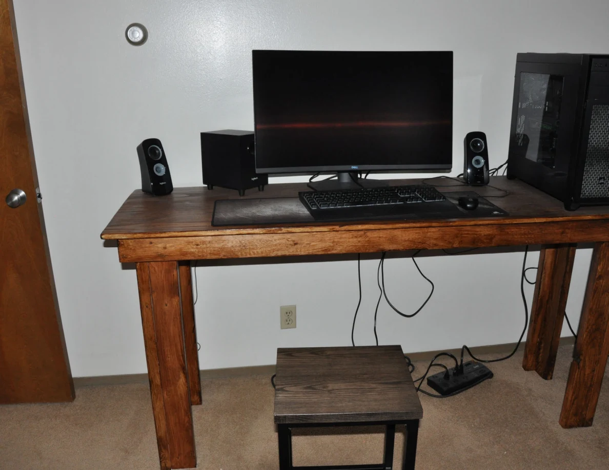 Computer workstation on a wooden desk with a black stool and cables underneath.