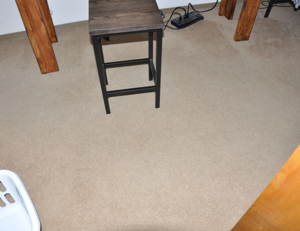 Black tall stool beneath a wooden desk, with a power strip and cords on carpet.