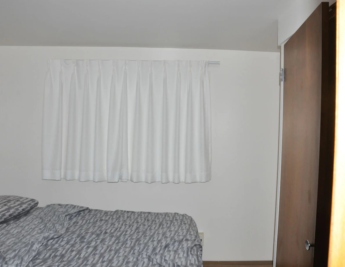 White curtains above a gray-covered bed beside a wooden closet door.