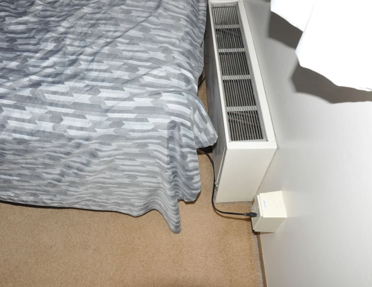 Bed edge beside a wall heater, curtains and a plugged-in cable.