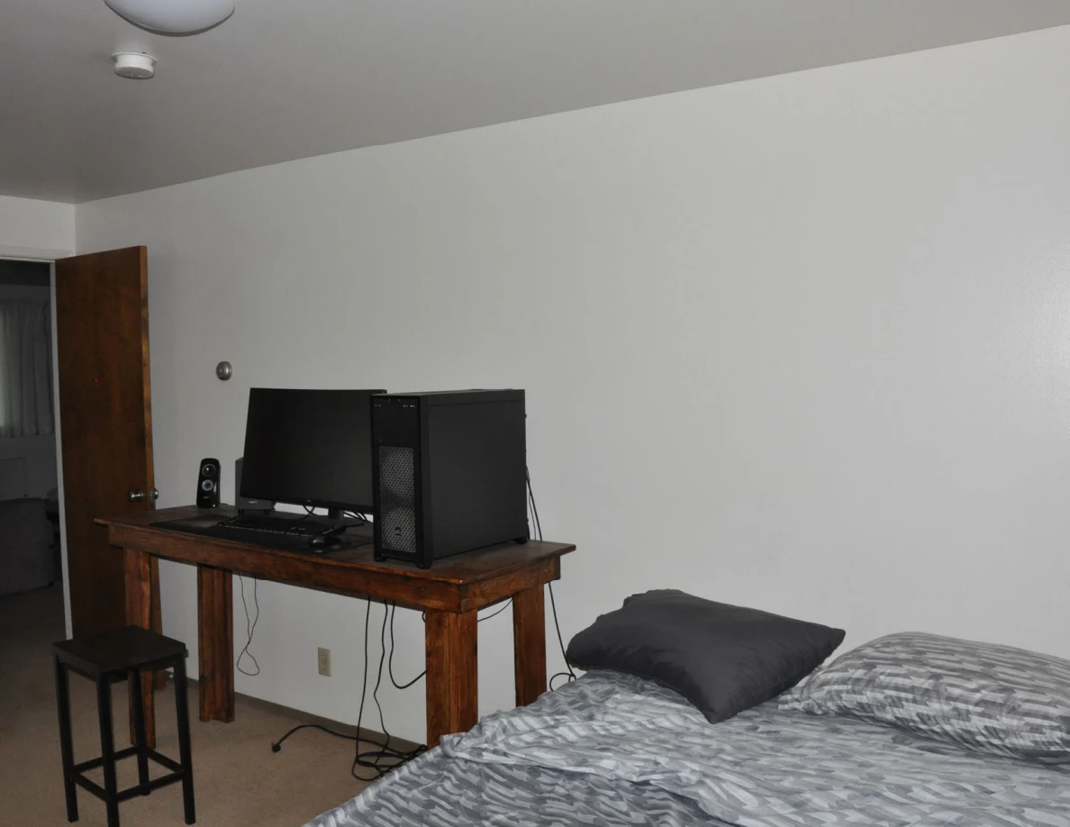 Bedroom with bed, wooden computer desk and stool under a ceiling light.