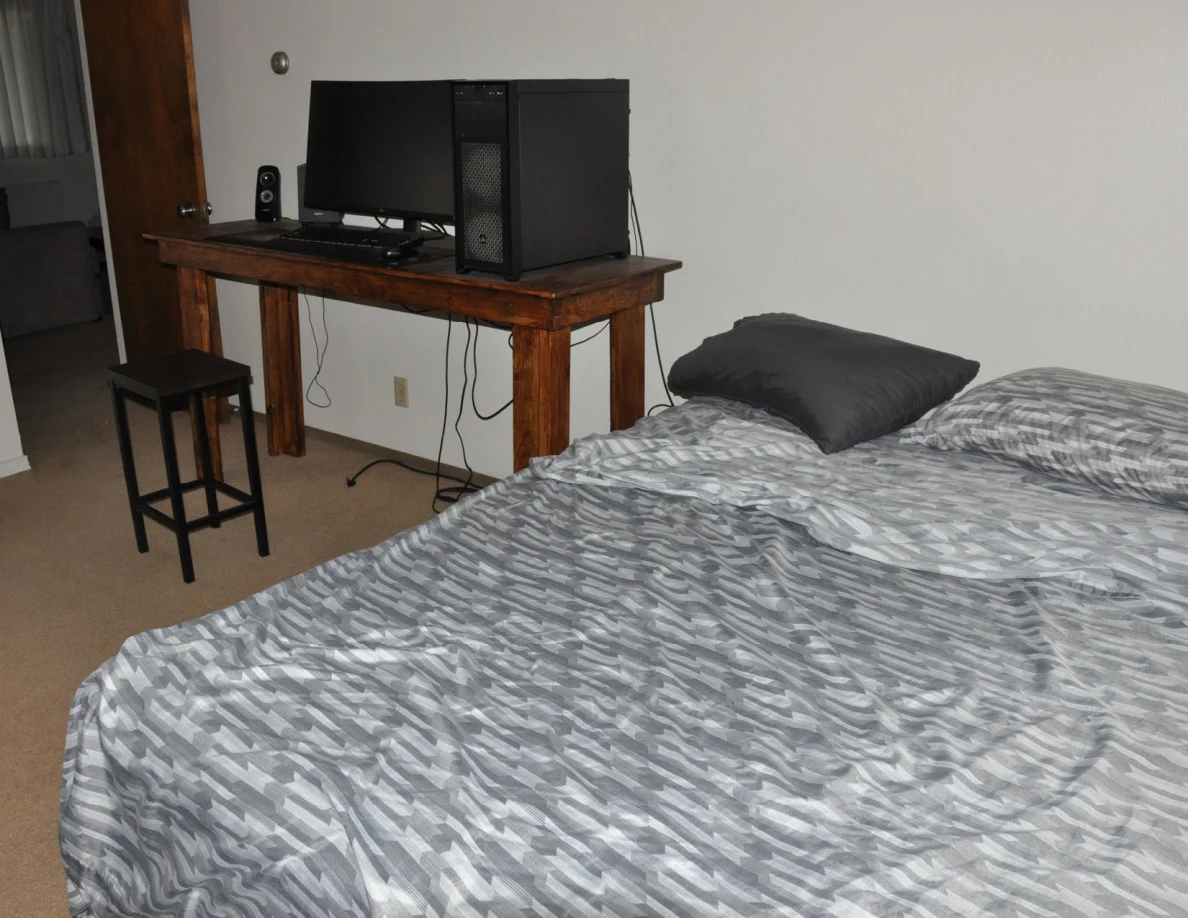 Gray patterned bedspread in the foreground with a computer workstation against the wall.