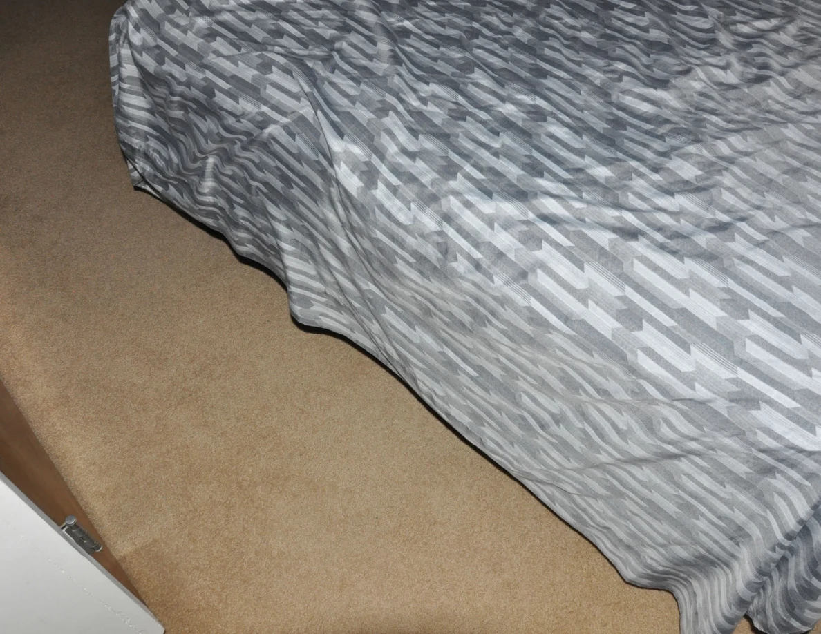 Close view of the side of a gray-patterned bedspread above beige carpet.