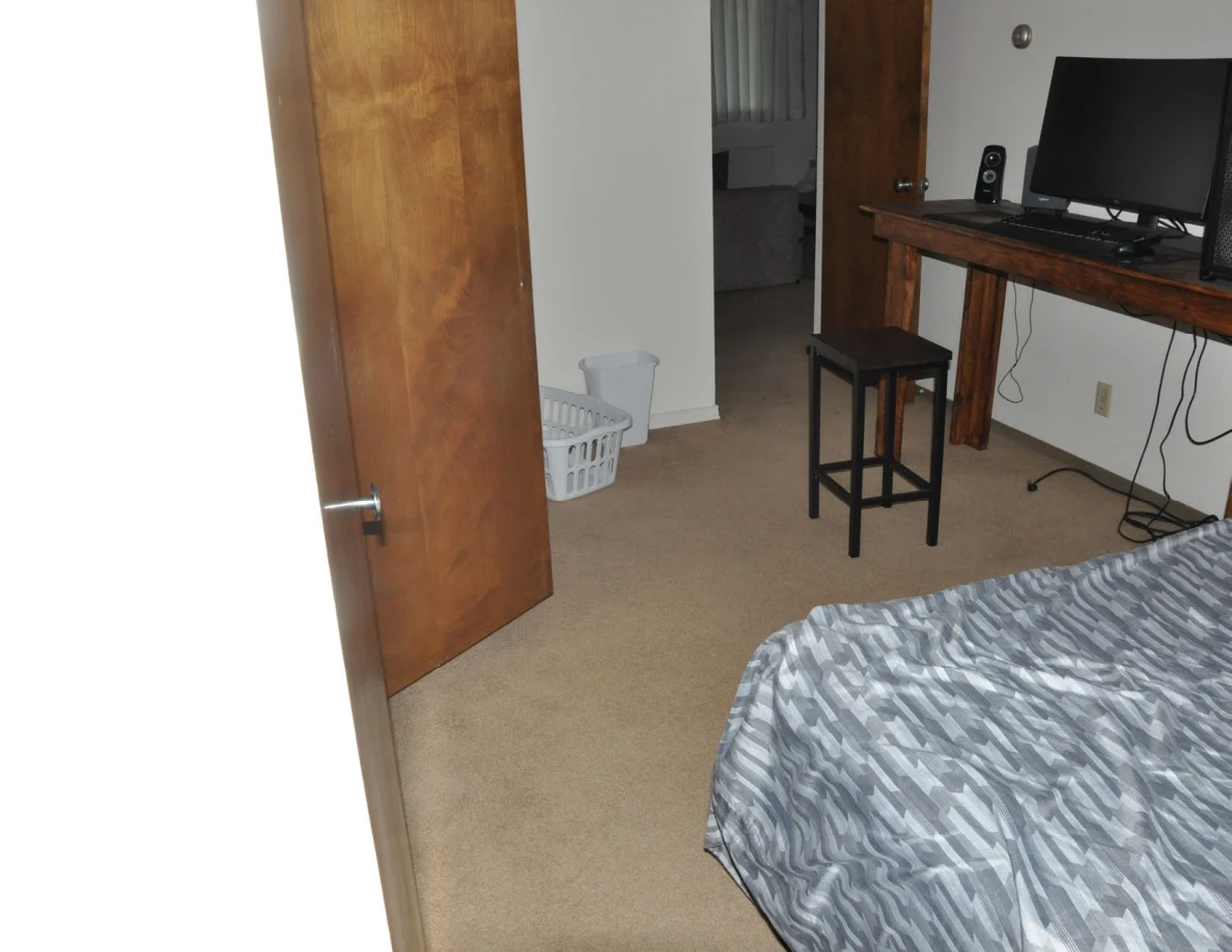 Bedroom with computer desk, stool, laundry basket and bed beside an open wooden door.