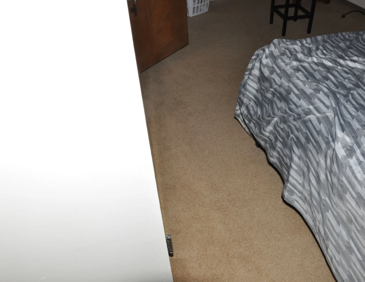 Narrow carpeted space along the side of a bed toward a stool and doorway.