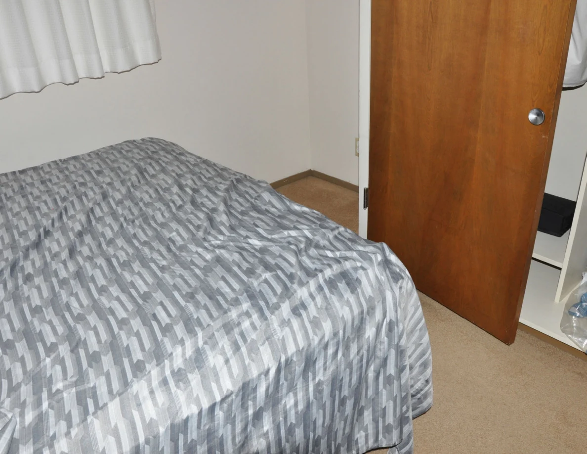 Foot of a gray-covered bed beside a wooden closet door.