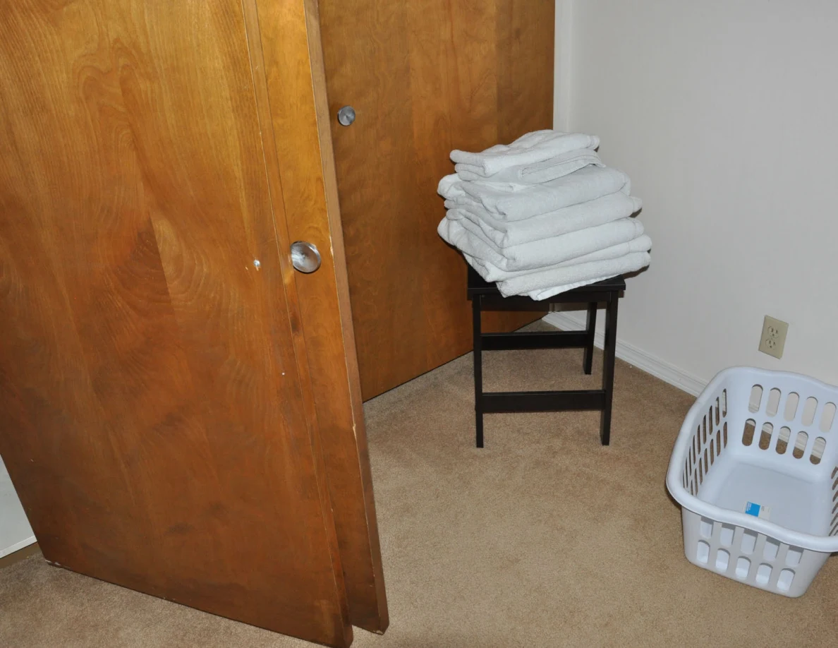 Stack of white towels on a black stool beside a laundry basket and wooden closet doors.