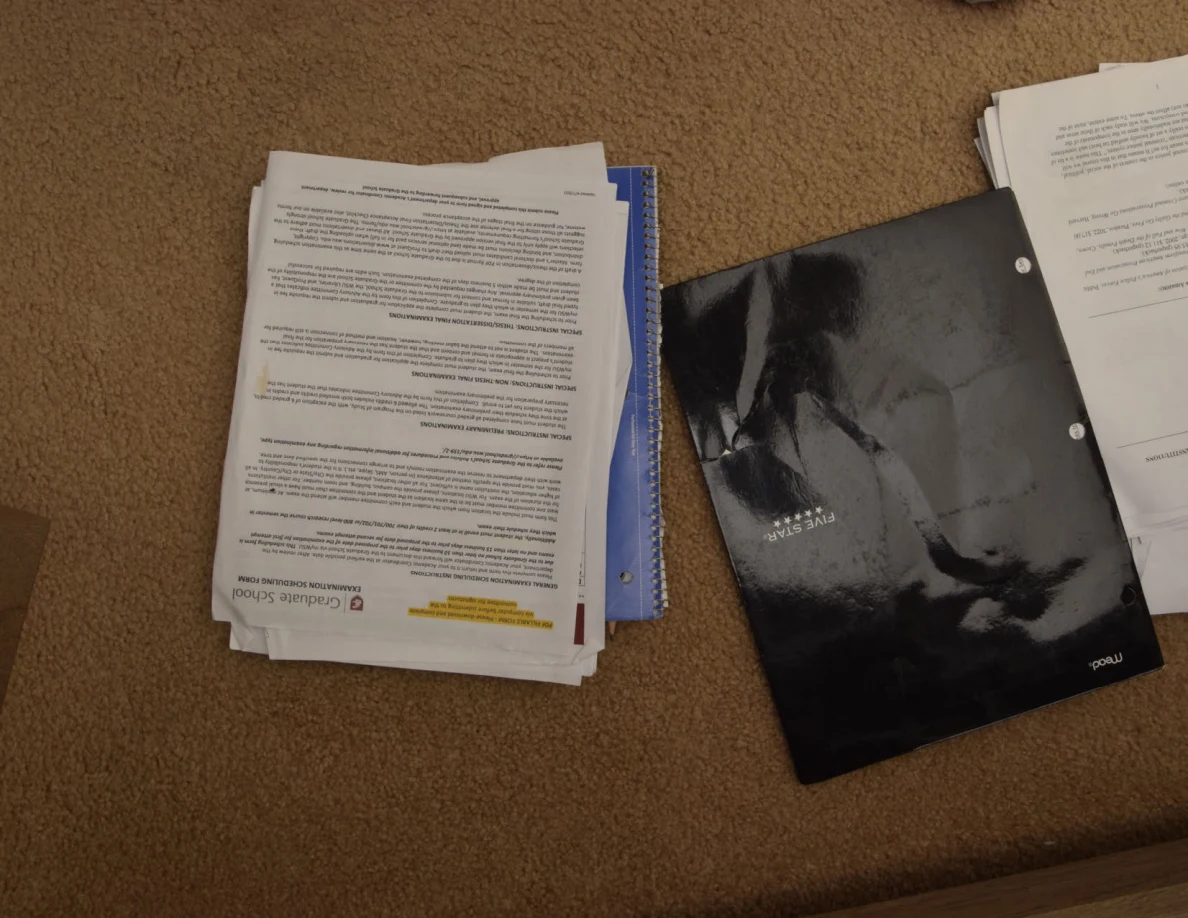 Stacks of printed graduate-school papers beside a black folder on carpet.