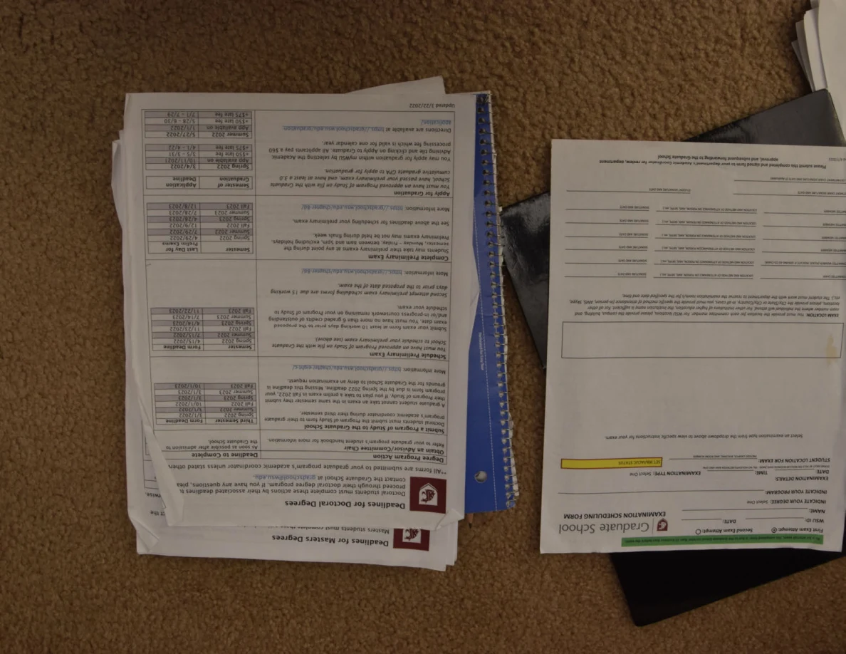 Printed graduate-school documents with tables laid over notebooks on carpet.