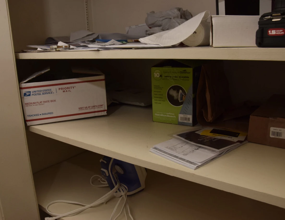 Lower storage shelves with mailing box, green package, paper stacks and an iron.