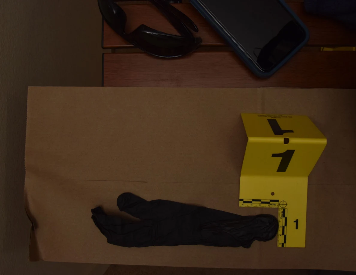 Black disposable glove on brown paper beside yellow marker 1; sunglasses and a phone sit beyond it.