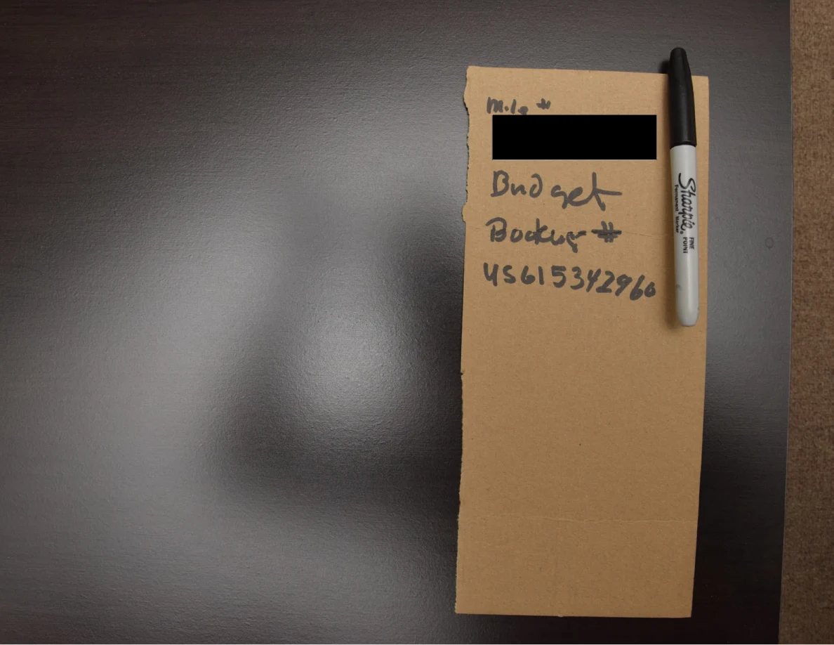 Handwritten note on brown cardboard beside a black marker pen, with one line redacted.