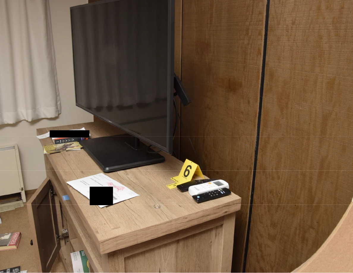 Television and remote controls on a wooden cabinet beside yellow marker 6, with papers partly redacted.