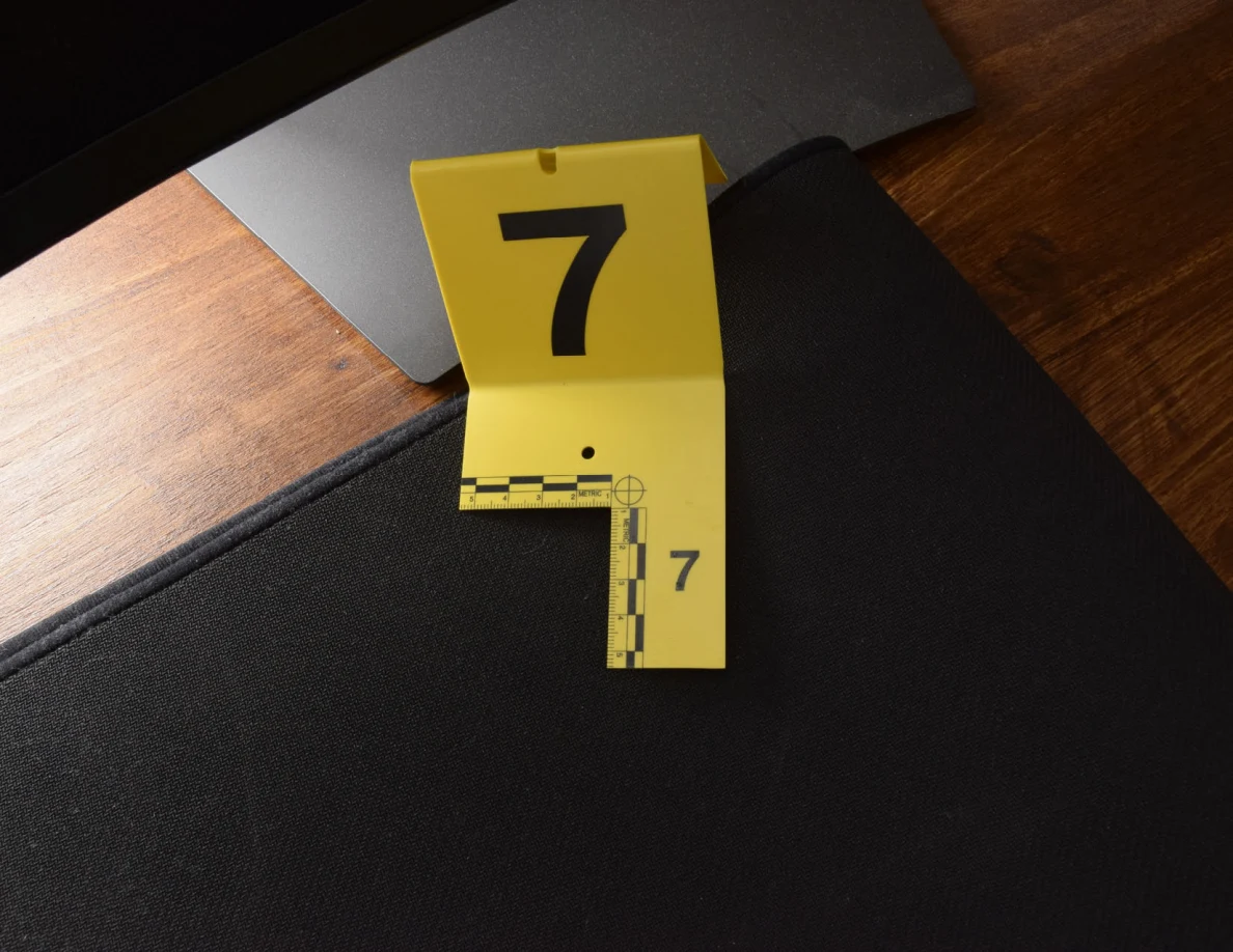 Yellow marker 7 on a black desk mat beside a monitor stand, under bright light.