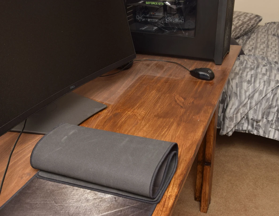 Computer desk with monitor, mouse, tower and a rolled-back desk mat beside a bed.
