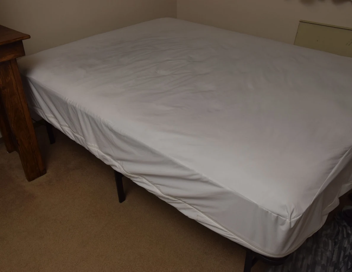 Bed covered by a plain white mattress cover beside a wooden desk.