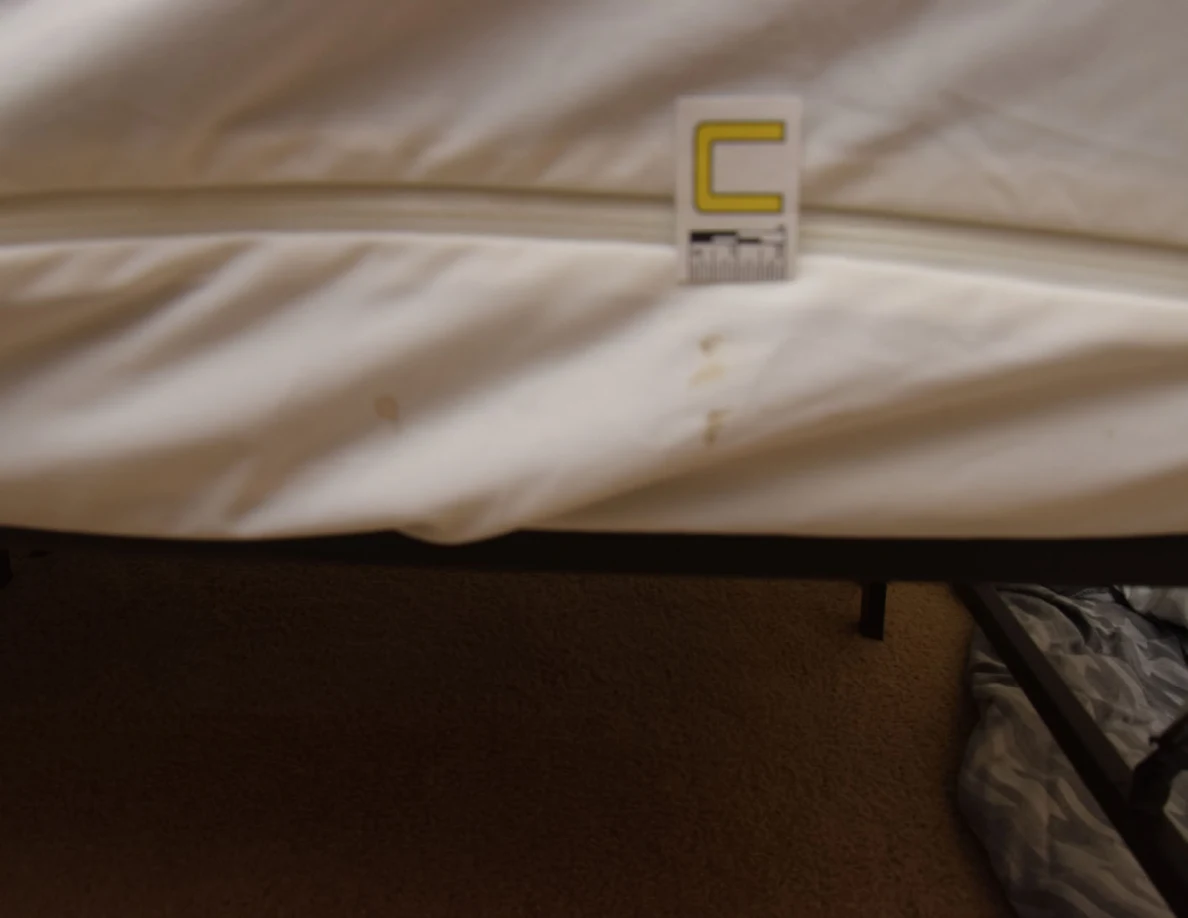 Close view of small tan marks near the seam of a white mattress cover, beside marker C.