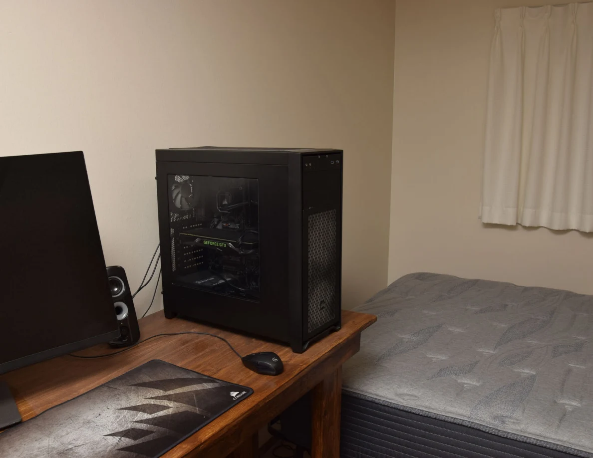 Computer tower and monitor on a desk beside an uncovered gray mattress.