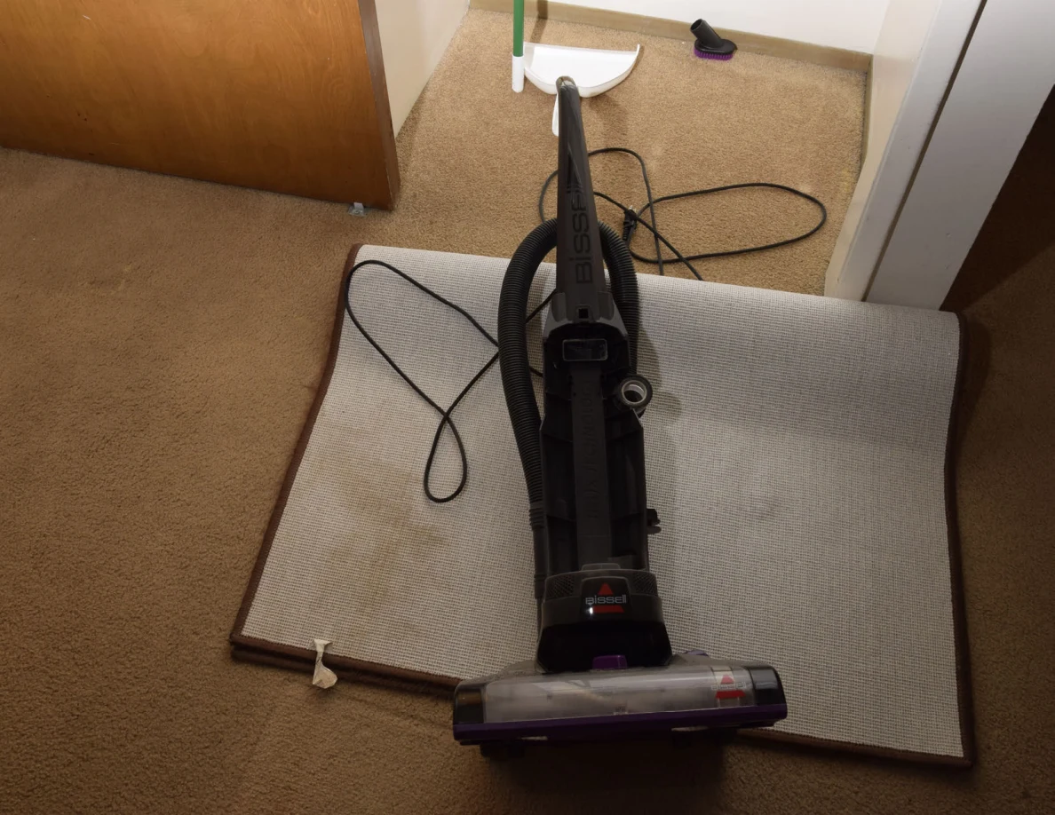 Upright vacuum laid flat on a rolled-back rug with its cord trailing across carpet.