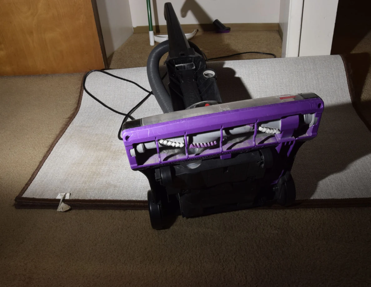 Underside of a vacuum cleaner showing purple base and brush roll over a folded-back rug.