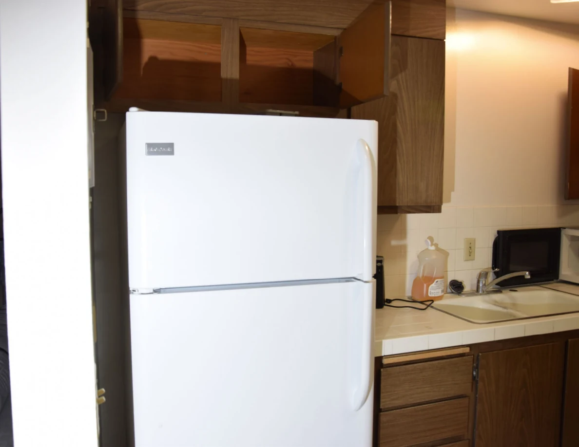 White refrigerator beneath open wooden cupboards beside a kitchen sink.
